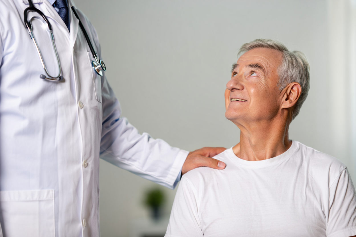 Врач в белом халате кладёт руку на плечо пожилому мужчине в белой футболке — поддержка при лечении алкоголизма в стационаре